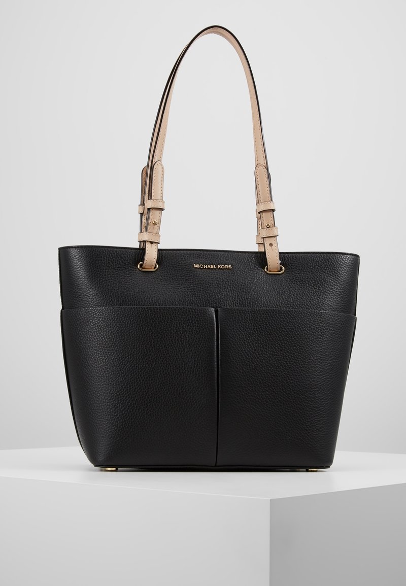 Musta tekstuurilla varustettu nahkatote-laukku, jossa on kaksi etutaskua ja beige nahkahihnat. Varustettu kultaisilla yksityiskohdilla ja logolla edessä.