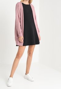 Ljust rosa, långärmad kofta över en svart, knälång klänning med figurnära livstycke. Vita sneakers fulländar outfiten.