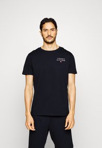 Zwarte katoenen t-shirt met korte mouwen, ronde halslijn, bevat een klein wit "TOMMY HILFIGER" logo aan de bovenste linkerzijde.