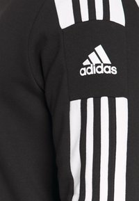 Svart hettegenser med hvite tre-striper på ermene og et adidas-logo på overarmen. Stoffet er en blanding av bomull.