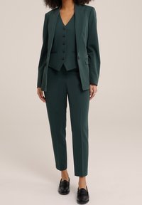 Costume ajusté vert foncé comprenant une veste à un bouton, un gilet assorti à cinq boutons et un pantalon droit. Des mocassins noirs complètent le look.