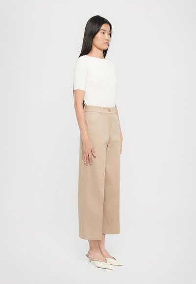 Beige weite Hosen, hochgeschnitten mit flachem Bund und Gürtelschlaufen. Dazu ein eng anliegendes weißes Rippenshirt und cremefarbene Slingback-Schuhe.