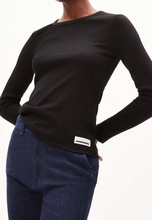 Persoon draagt een strak zwart shirt met lange mouwen met een klein wit ARMEDANGELS-logo label, gecombineerd met donkerblauwe spijkerbroek.