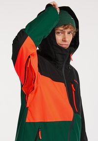 Kleurrijke jas met zwarte, groene en oranje panelen. Heeft een capuchon, ritszakken en een sportief ontwerp met een synthetische textuur.