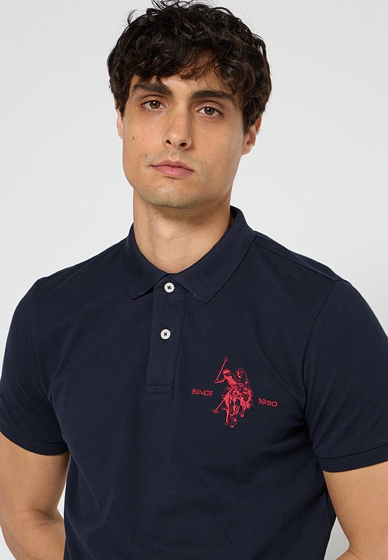 Polo en coton mélangé de couleur marine avec des manches courtes, patte de boutonnage à deux boutons et un logo rouge brodé représentant un joueur de polo avec le texte "DEPUIS 1990".