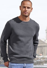 Grau strukturierter Strickpullover mit Rundhalsausschnitt, der diagonale Rippmuster und gerippte Bündchen aufweist. Getragen mit blauen Jeans.