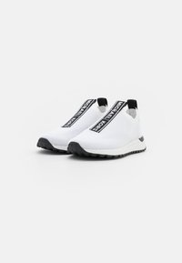 Bílé pletené tenisky s černou gumovou podrážkou, s výrazným černým páskem označeným "MICHAEL KORS" přes svršek.