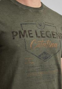 T-shirt en coton vert olive foncé avec une impression graphique en bleu foncé et orange. Col rond, manches courtes et design décontracté.