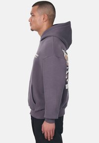 Grauer Kapuzenpulli mit einer Vordertasche, grafischen Seitennähten und großer Kapuze. Der Stoff wirkt strukturiert und weist gerippte Bündchen sowie einen gerippten Saum auf.