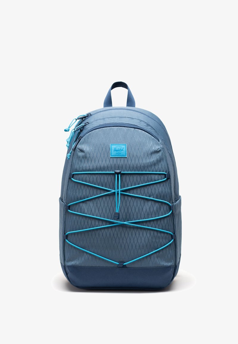Sac à dos bleu avec devant à motif losange, détail de cordon élastique bleu clair, poches zippées et poignée sur le dessus, sur fond blanc.