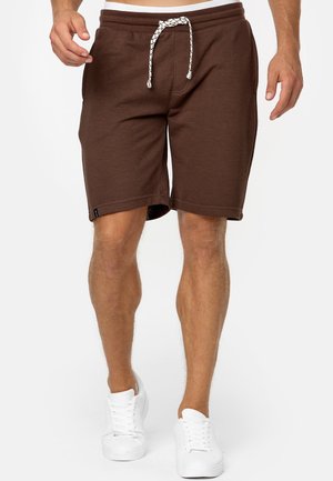Gambe maschili che indossano pantaloncini marroni con coulisse e sneaker bianche su uno sfondo chiaro e uniforme.