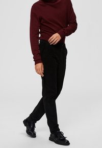 Schwarze Cordhosen mit schmal zulaufendem Schnitt und strukturierter Oberfläche. Kombiniert mit einem langärmligen Oberteil in Bordeauxrot und schwarzen Schuhen.