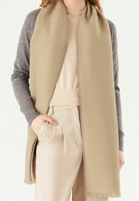 Écharpe en mélange de laine beige avec des bords frangés, portée ouverte sur un haut gris clair et un pantalon beige. Les manches grises texturées ajoutent du contraste.