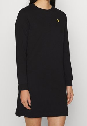 Kobieta ubrana w czarną sukienkę z długim rękawem z małym żółtym logo ptaka na górnej lewej części klatki piersiowej.