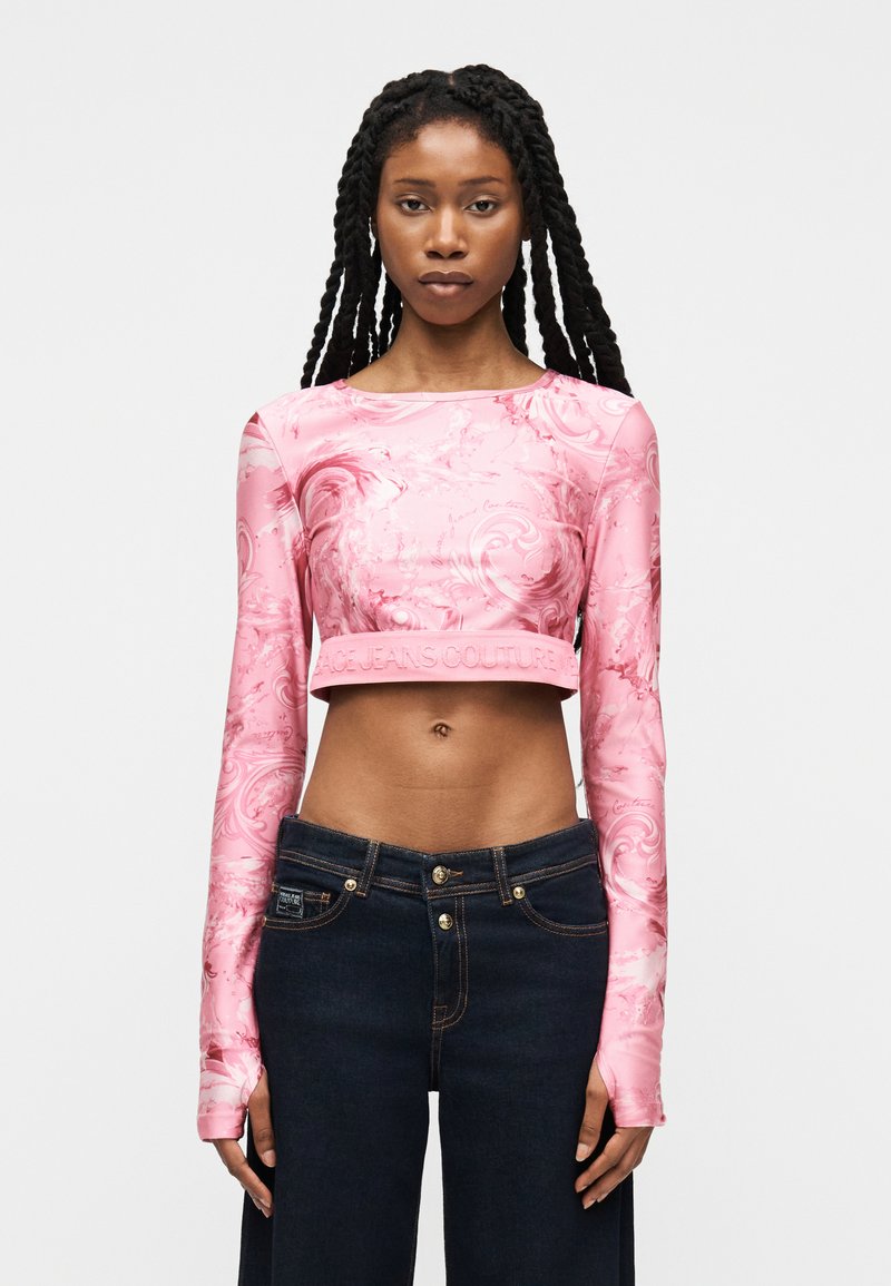 Γυναίκα με μακριά πλεξούδα, που φοράει ροζ crop top με μακριά μανίκια και σκοτεινά μπλε ψηλόμεσα τζιν, κοιτάζοντας προς τα εμπρός.