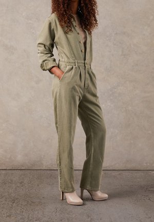 Femme portant une combinaison verte olive boutonnée, manches retroussées et bottes à talons beiges, les mains dans les poches, debout sur un sol texturé.