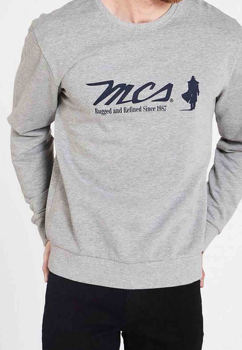 Homme portant un sweat-shirt gris clair avec le texte « MCS Rugged and Refined Since 1987 » et une silhouette d'une personne portant un chapeau et un manteau sur la poitrine.