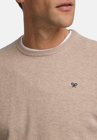 Suéter de punto beige con cuello redondo acanalado y pequeño bordado oscuro. La tela parece suave con una textura lisa.