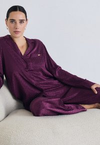 Nainen, jolla on tummat hiukset ja siniset silmät, makaa vaalealla sohvallaan, yllään teksturoitu syvän purppuranvärinen pitkähihainen pyjamapuku ja kultaiset renkaat korvissa.