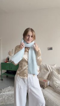 Cachecol azul-claro canelado enrolado ao pescoço, feito de tecido texturizado. Modelo veste uma blusa bege de cetim e calças brancas largas.