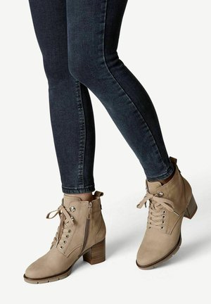 Botines beige de ante, con tacón cuadrado, cordones en la parte delantera y cremallera lateral; textura suave, punta redonda y detalles decorativos.