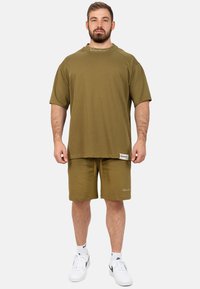 T-shirt in cotone verde oliva con maniche corte e una vestibilità ampia, abbinato a pantaloni corti coordinati, presenta un logo e un dettaglio dell'etichetta. Sneakers bianche.