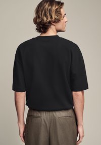 Zwarte kortekous T-shirt van zachte stof, met een relaxed fit en een ronde halslijn, gedragen met lichtbruine broeken.