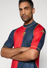 Krótka koszulka sportowa z czerwonymi i granatowymi panelami, złotymi akcentami oraz emblematem lwa na piersi i rękawie, wykonana z gładkiej tkaniny.