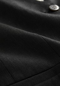 Gros plan sur un tissu noir avec des coutures visibles et un bouton foncé sur un vêtement, mettant en valeur la texture et les coutures.