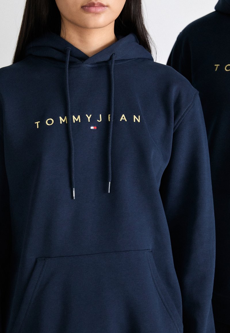 Sweat à capuche bleu marine en tissu doux avec poche frontale, arborant un logo doré "TOMMYJEAN" et un petit accent de logo en rouge et blanc.