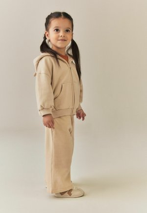 Ragazza giovane con capelli scuri intrecciati che indossa una felpa con cappuccio beige, pantaloni abbinati e scarpe beige, in piedi davanti a uno sfondo semplice.