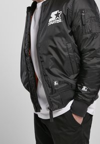 Bomber nera con zip sul davanti e polsini a costine. Presenta il logo bianco "STARTER", tasche laterali e una piccola etichetta del marchio sull'orlo.