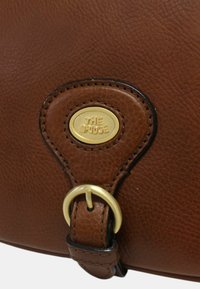 Dettaglio dell'angolo di una borsa in pelle marrone con una placca ovale dorata con la scritta "The Bridge" e un anello metallico dorato attaccato a una tracolla in pelle.
