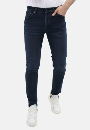 Donkerblauwe slim-fit jeans met vijf zakken, zilveren hardware en een middelhoge taille, gecombineerd met witte sneakers. Gladde denimtextuur.