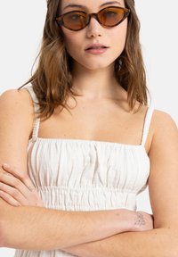 Witte, gestructureerde zomerjurk met smalle bandjes en een geraffineerde taille. Bruin getinte cat-eye zonnebril. Model met lichtbruin haar, met gekruiste armen.