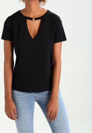 Vrouw die een zwart shirt met korte mouwen draagt met een kleine metalen ringdetail bij de halslijn, gecombineerd met lichtblauwe spijkerbroek.