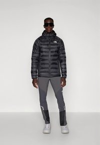 Črni puhovka z zadrgo spredaj in kapuco, kombinirana s sivimi hlačami z belimi poudarki. Dodatki vključujejo temna sončna očala in bele superge.