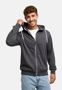 Glimlachende man die de capuchon van een donkergrijze rits-hoodie over een wit shirt aanpast, gekleed in lichtblauwe jeans, tegen een effen achtergrond.
