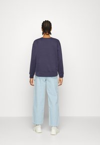 Felpa navy con polsini e orlo a coste, abbinata a jeans a gamba larga di colore azzurro chiaro. La modella è di spalle, su uno sfondo bianco semplice.