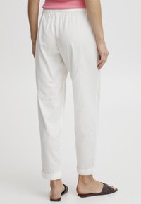 Pantalon en lin blanc avec une taille élastique, des poignets retroussés et une coupe décontractée. Le modèle porte un haut rose et des sandales brunes.