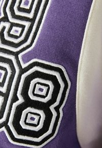 Lilla tekstil med store, sorte og hvide broderede tal '9' og '8', med geometriske mønstre og en glat tekstur.
