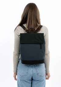 Sac à dos noir et marine avec un rabat, en matériau texturé, finition lisse, et une poignée supérieure. Bretelles réglables et détails matériels minimalistes.