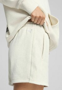 Pantalones cortos de color beige claro con una textura suave, que cuentan con una banda elástica en la cintura, bolsillos laterales y un pequeño logo metálico en el dobladillo.