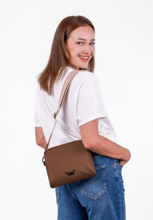 Hnědá crossbody taška s hladkou teksturou, zipovým uzávěrem a tkaným popruhem. Černý logo akcent na přední straně. Nosená ležérně s džínami.