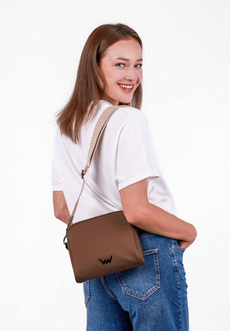 Braune Umhängetasche mit glatter Textur, Reißverschluss und gewebtem Riemen. Schwarzes Logo-Highlight auf der Vorderseite. Lässig zu Jeans getragen.