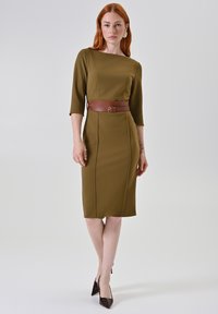 Olivengroene nauwsluitende jurk met een boothals, driekwart mouwen, een bruine riem in de taille en een zoom tot op de knie. Gladde textuur.