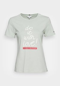 Camiseta de manga corta gris claro con el texto "¡hazlo con amor!" en blanco y "Tommy Hilfiger" en rojo debajo, pequeño logotipo en la manga izquierda.
