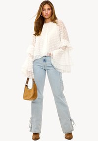 Blouse blanche à volants avec des pois texturés, associée à un jean taille haute bleu clair. Le modèle porte un sac à main beige et des bottes marron.
