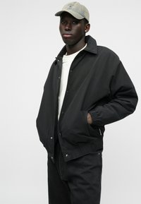 Melnā bumbiera jaka ar spiedpogām, sānu kabatām un elastīgām piedurknēm. Uzvilkta pār baltu kreklu, kopā ar melnajām džinsām un pelēko cepuri.