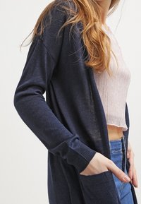Dlouhý navy cardigan vyrobený z měkké pleteniny, s volným střihem, žebrovanými manžetami a bočními kapsami, vrstvený přes světle růžový top.